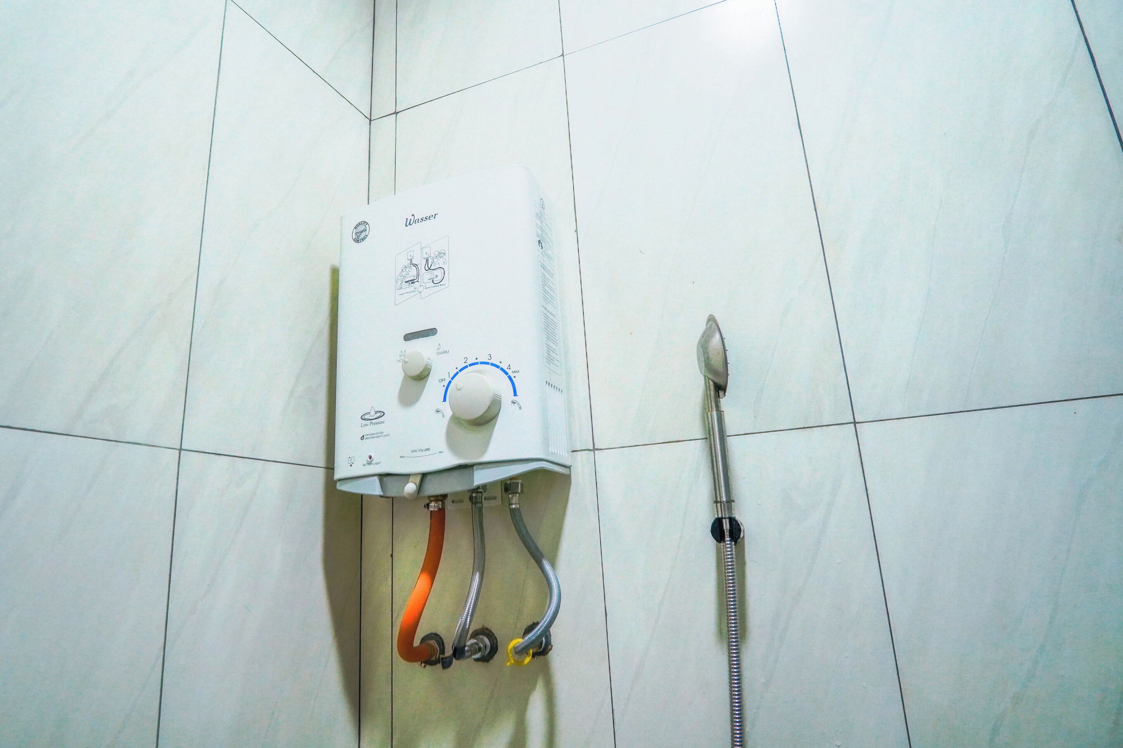 A water heater with a control knob, two hoses, and a showerhead mounted on a tiled wall.