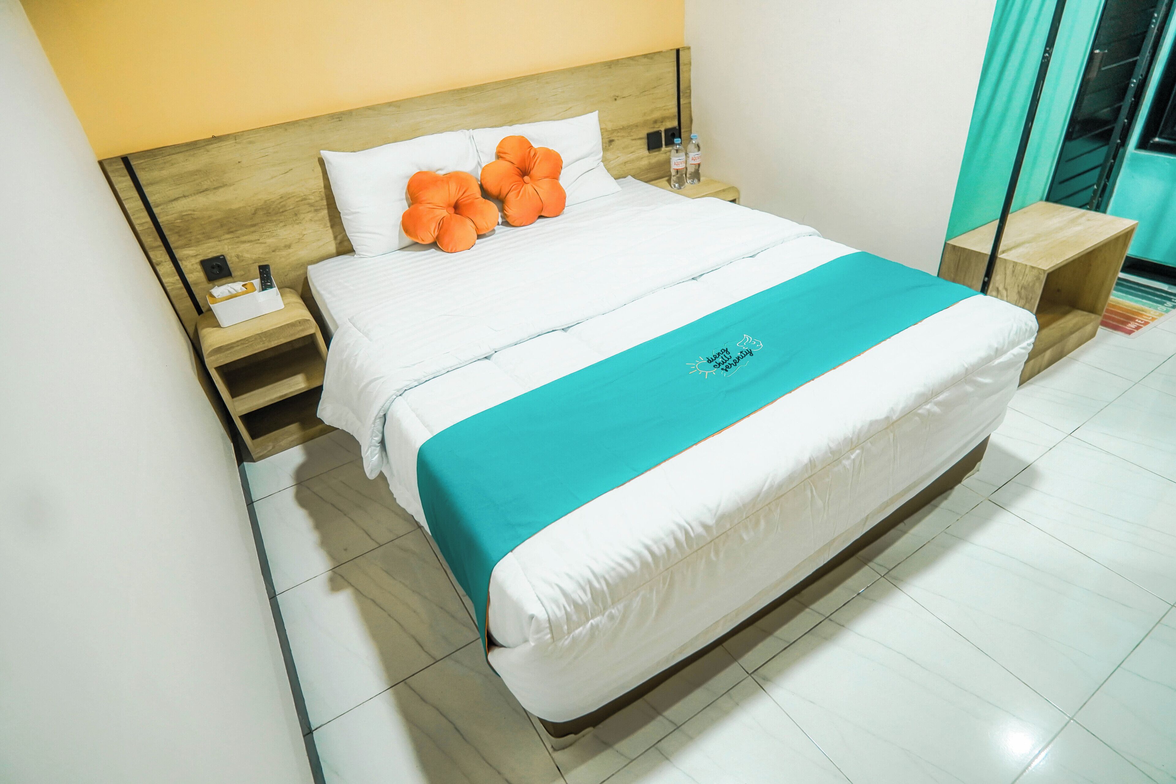Una cama bien hecha con colcha blanca y azul verdoso, cabecera de madera y mesita de noche con una botella y un control remoto.