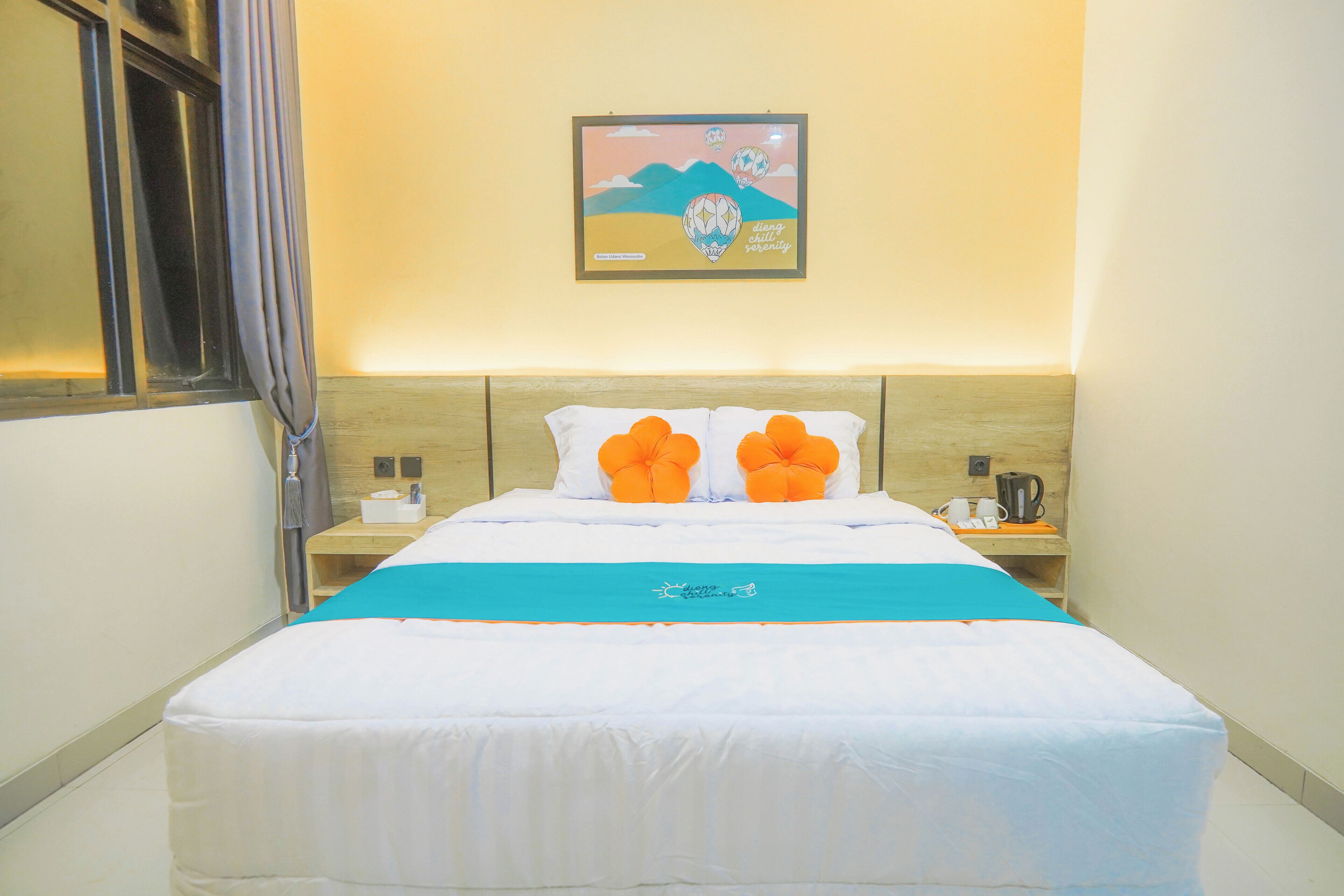 Habitació d'hotel amb un llit, una tauleta de nit amb un telèfon i una tetera, un quadre penjat a la paret i una finestra amb cortines.