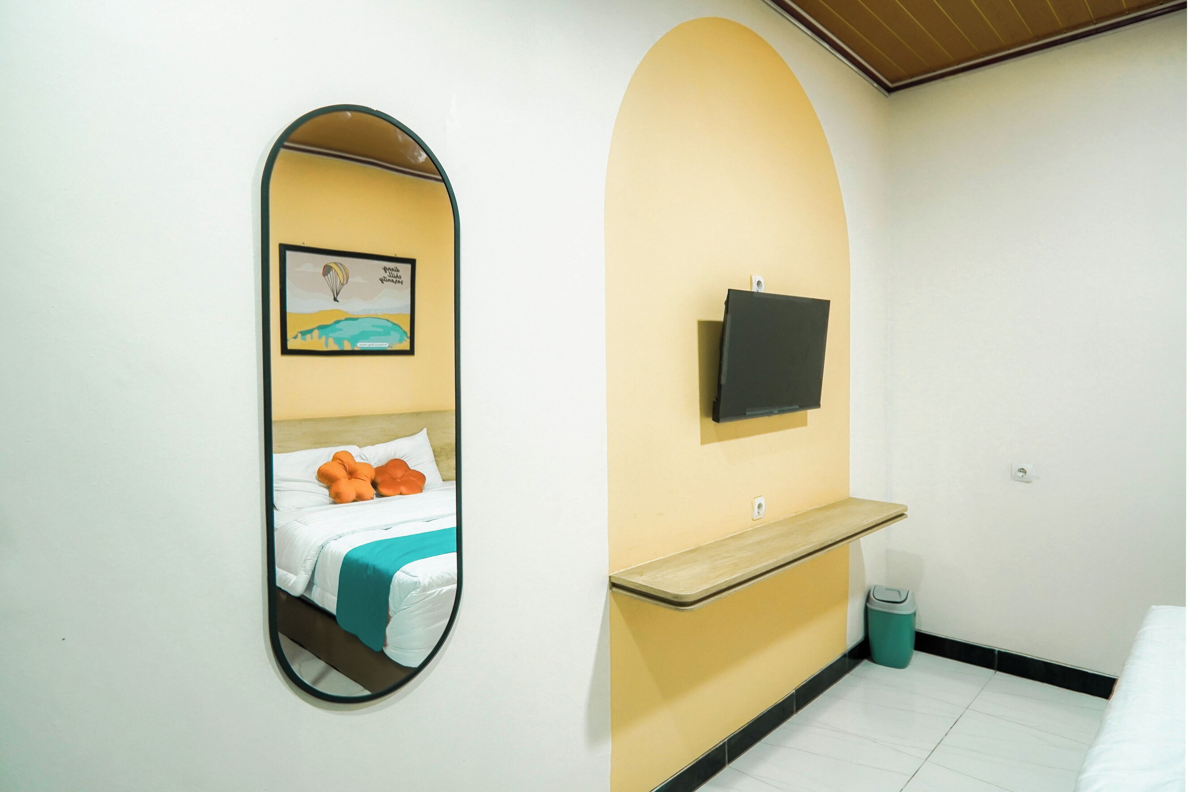A hotel room with a bed, a mirror, a mounted TV, a small shelf, and a trash bin.