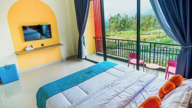 A hotel room with a bed, a TV mounted on a yellow wall, a balcony with chairs, and a view of greenery. - INSTAY Hotel Dieng Chill Serenity (Garung)