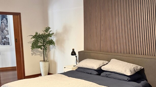 Quarto moderno com uma cama grande, uma planta na mesa de cabeceira e uma fotografia de um horizonte urbano na parede.