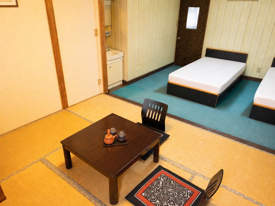 Una camera d'albergo con due letti, un piccolo tavolo con teiera, una sedia, un lavandino e uno specchio.