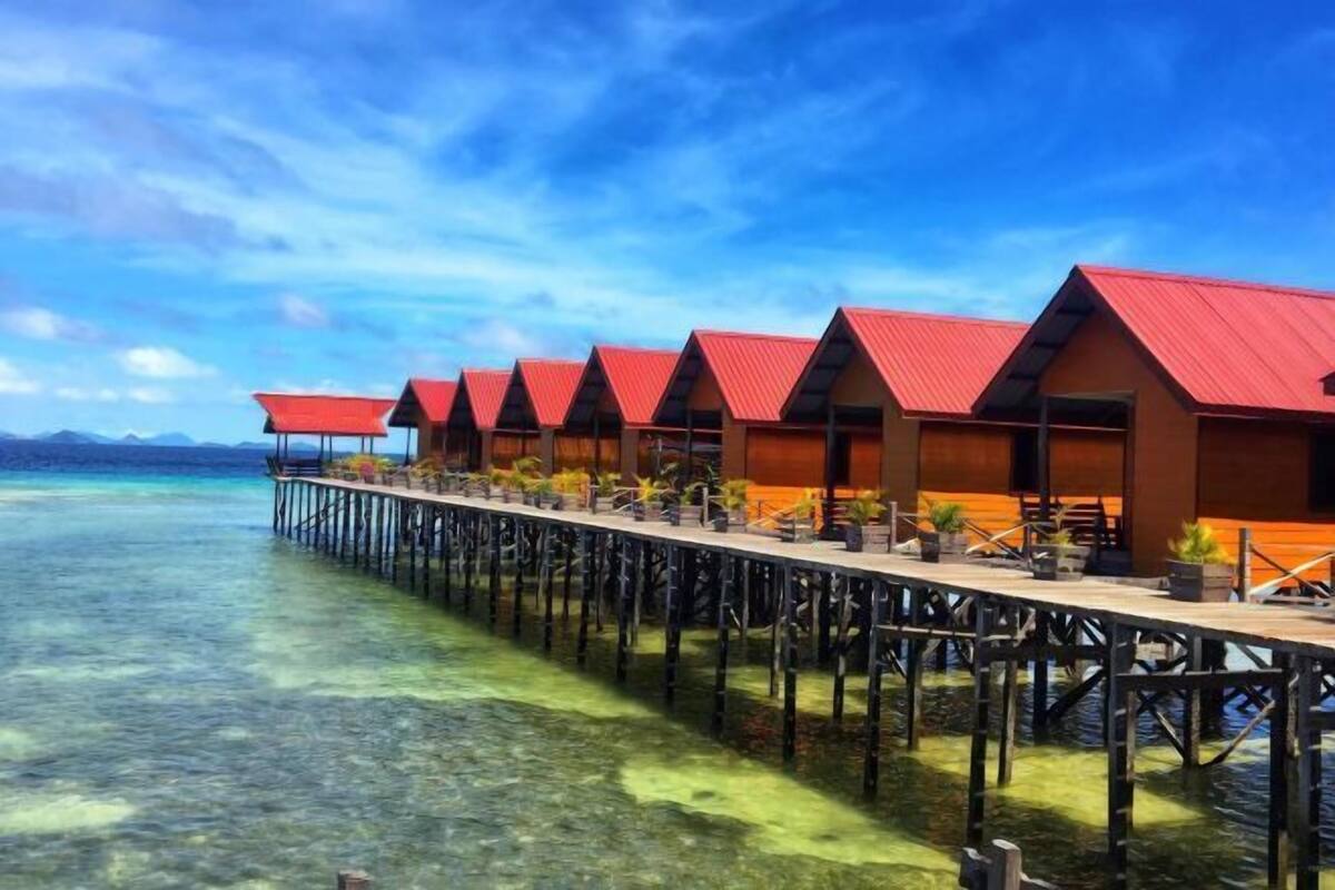 A row of overwater bungalows with red roofs, connected by a wooden walkway, overlooking clear turquoise water.