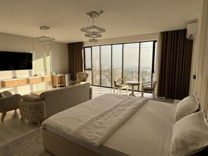 Ein Hotelzimmer mit einem großen Bett, einem Schreibtisch mit Flachbildfernseher, einem Sessel, einem kleinen Tisch und Blick auf die Stadt durch große Fenster.