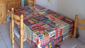 Ein Essbereich mit einem Tisch, der mit alten Werbeplakaten bedeckt ist, Holzstühle, einem weißen Drahtkorb und einem Holzschrank mit Glastüren.