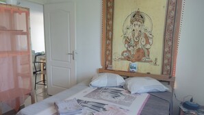 Una camera da letto con un letto, cuscini, una coperta, un arazzo a parete, una porta e un tavolino con una sedia.