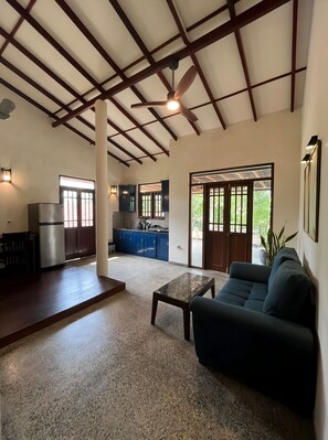 A spacious living area with a ceiling fan, a kitchen in the background, and a view through a glass door. - Sereniqe Heaven: Experience the Difference of True Serenity (Hikkaduwa)