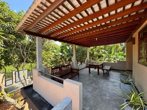 A covered outdoor seating area with a table and benches, surrounded by lush greenery.