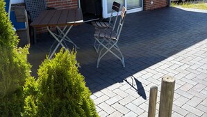 A building with a brick facade, a sliding glass door entrance, a table and chairs outside, and a parked car in the background. - Apartment Kleine Auszeit with Terrace and Wi-Fi (Dagebüll)