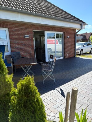 Ein Gebäude mit Ziegelgliederung, einer Schiebeglastür als Eingang, einem Tisch mit Stühlen draußen und einem parkenden Auto im Hintergrund.