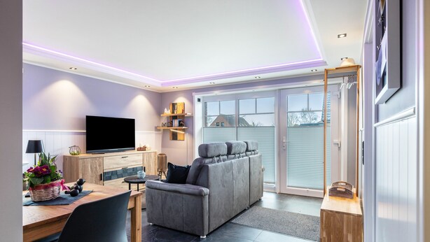 Una sala de estar moderna con un sofá, un televisor de pantalla plana, una mesa de comedor con sillas y una zona de cocina con ventanal.