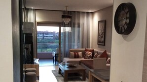 A modern living room with a sofa, coffee table, and dining table. There is a large window with curtains, a chandelier, and a clock on the wall. - Apartment Prestigia Jade pool and garden view (Marrakech)