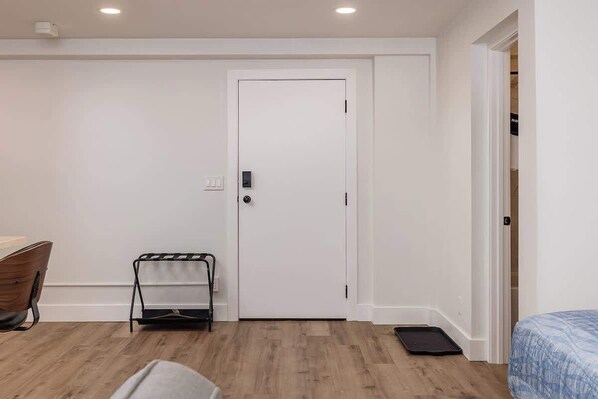 Una habitación con una puerta blanca, una mesita con una silla, una cama con colcha azul y suelo de madera.