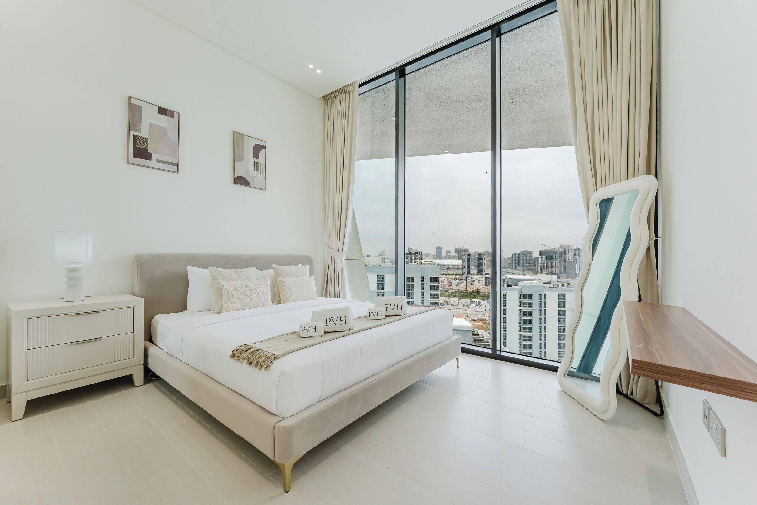 Une chambre moderne avec un grand lit, une table de chevet, un miroir et une vue sur la ville grâce à de grandes fenêtres.