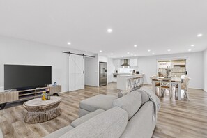 Ein modernes Wohnzimmer mit Flachbildfernseher, einem grauen Sofa, einem Korbtisch und einer Küche im Hintergrund.