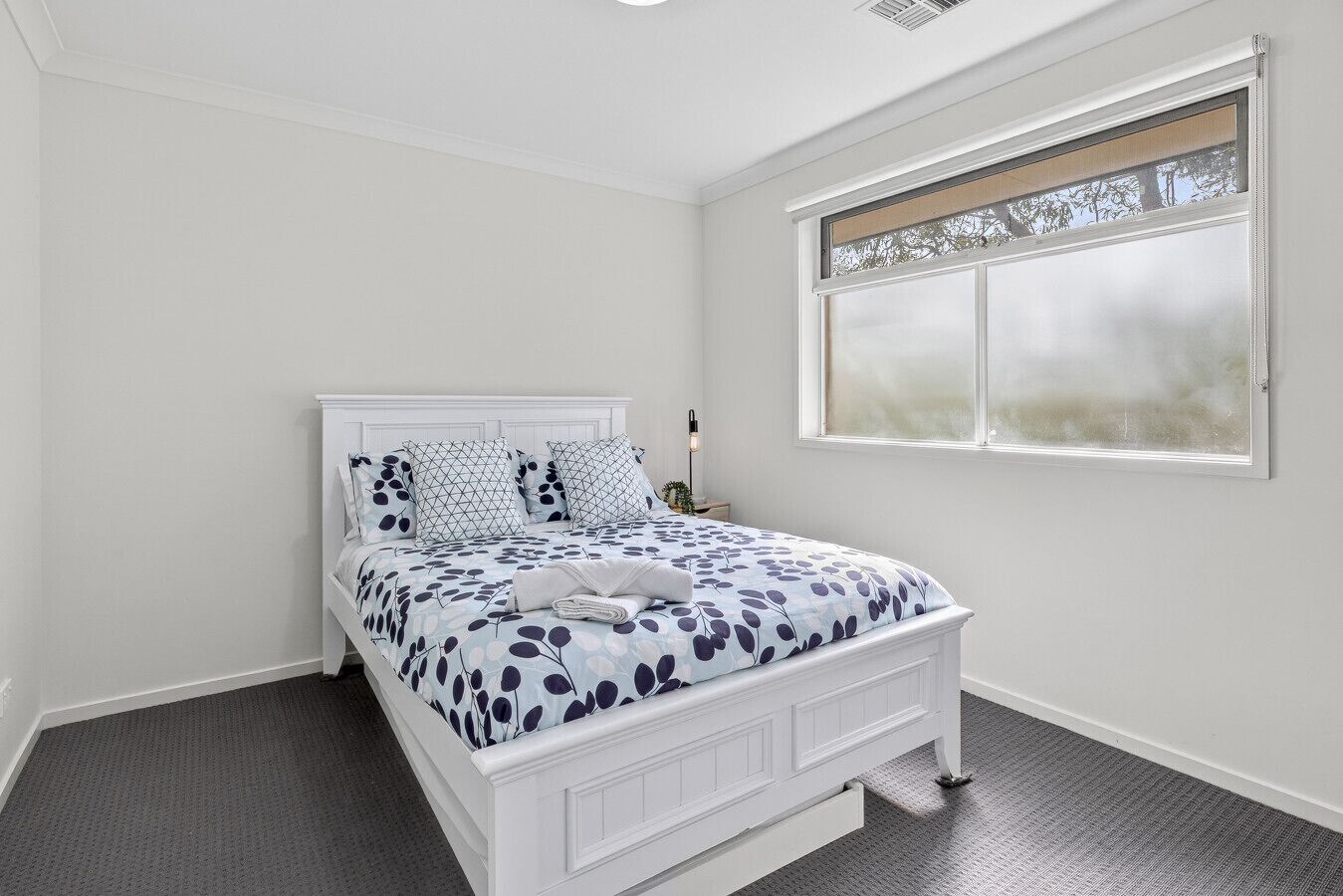 A bedroom with a white bed, a window with a view, and a nightstand with a lamp.