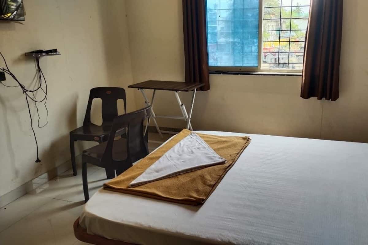 Un letto singolo con materasso bianco e coperta marrone, un tavolino, una sedia e una finestra con tende.