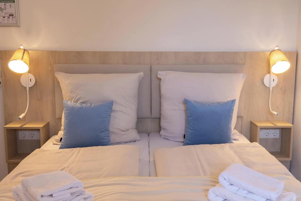 A double bed with white linens, two blue pillows, and two bedside tables with lamps.