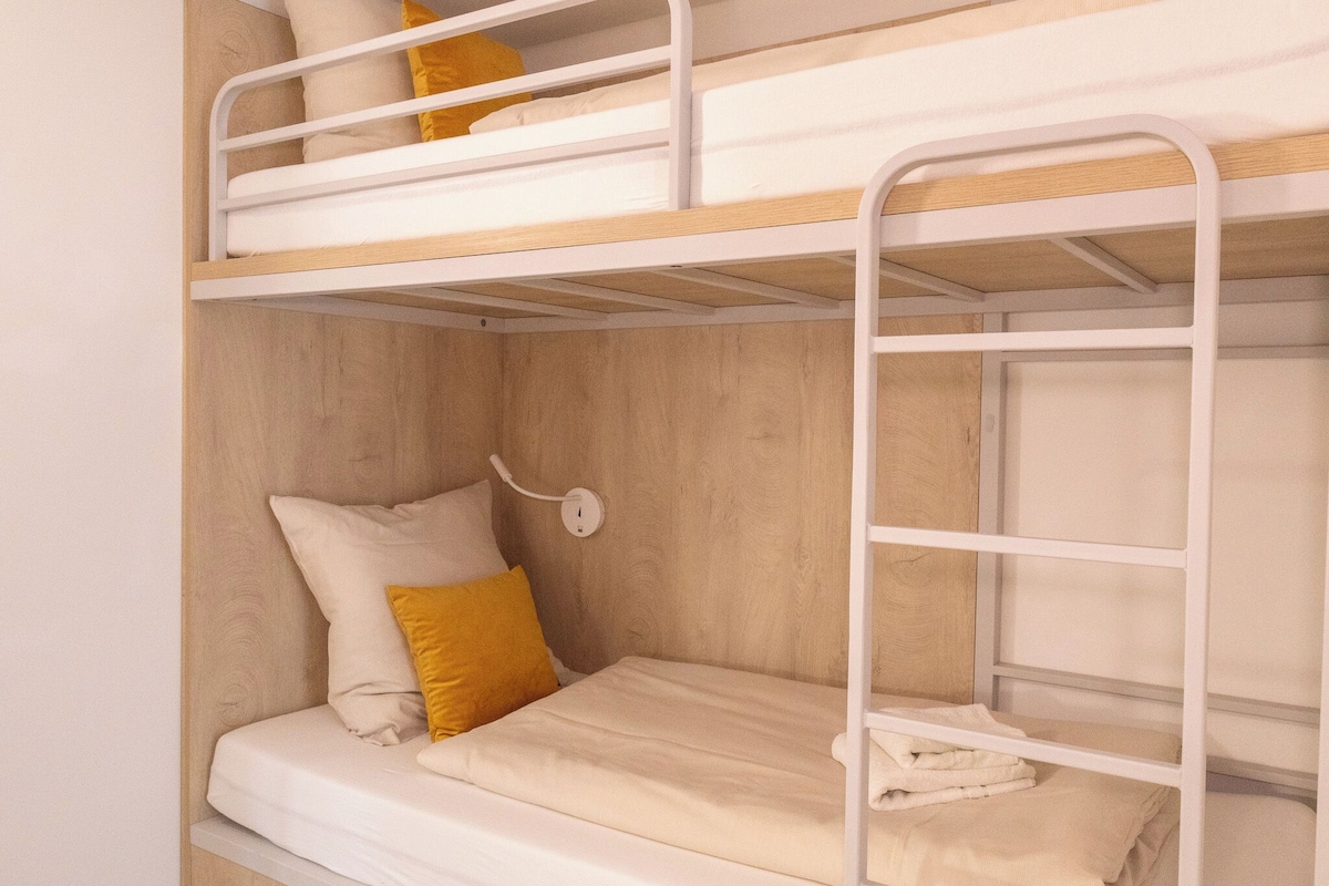 Dormitorio con literas, cabecera de madera, pared blanca y mesita de noche con lámpara.