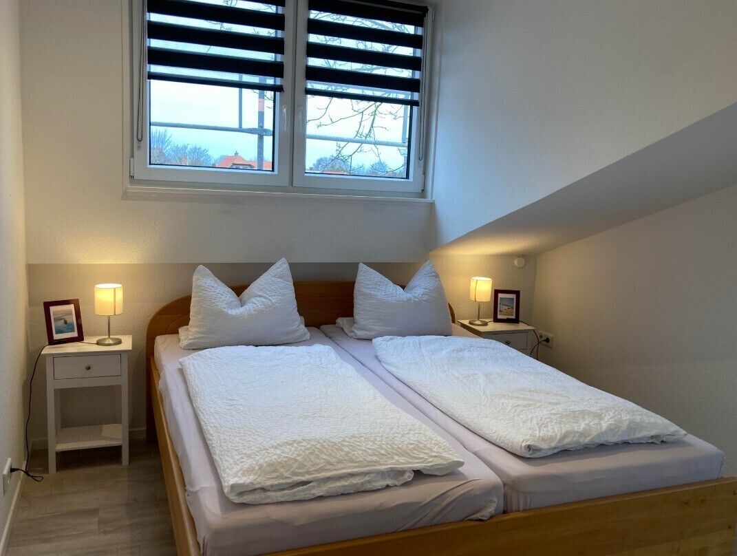 Ein Doppelbett mit weißer Bettwäsche, zwei Nachttische mit Lampen, ein Fenster mit Jalousien und eine schräge Decke.