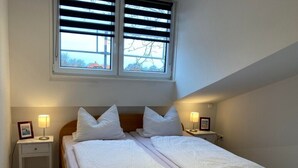 A twin bed with white bedding, two bedside tables with lamps, a window with blinds, and a sloped ceiling. - Cozy vacation apartment near the beach & town center (Langeoog)