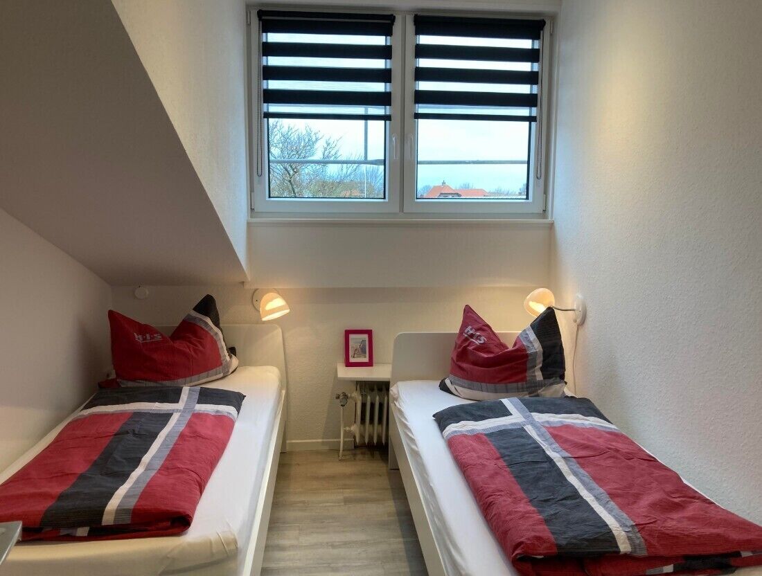 A small room with two single beds, each with a red and grey blanket, a window with blinds, a radiator, and a small table with a picture frame.