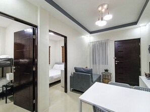 A hotel room with a bed, a desk, a chair, a small table, a potted plant, and a window with curtains. - Manzil Anilao Unit 1 - Ground Floor 1st Room (Mabini)