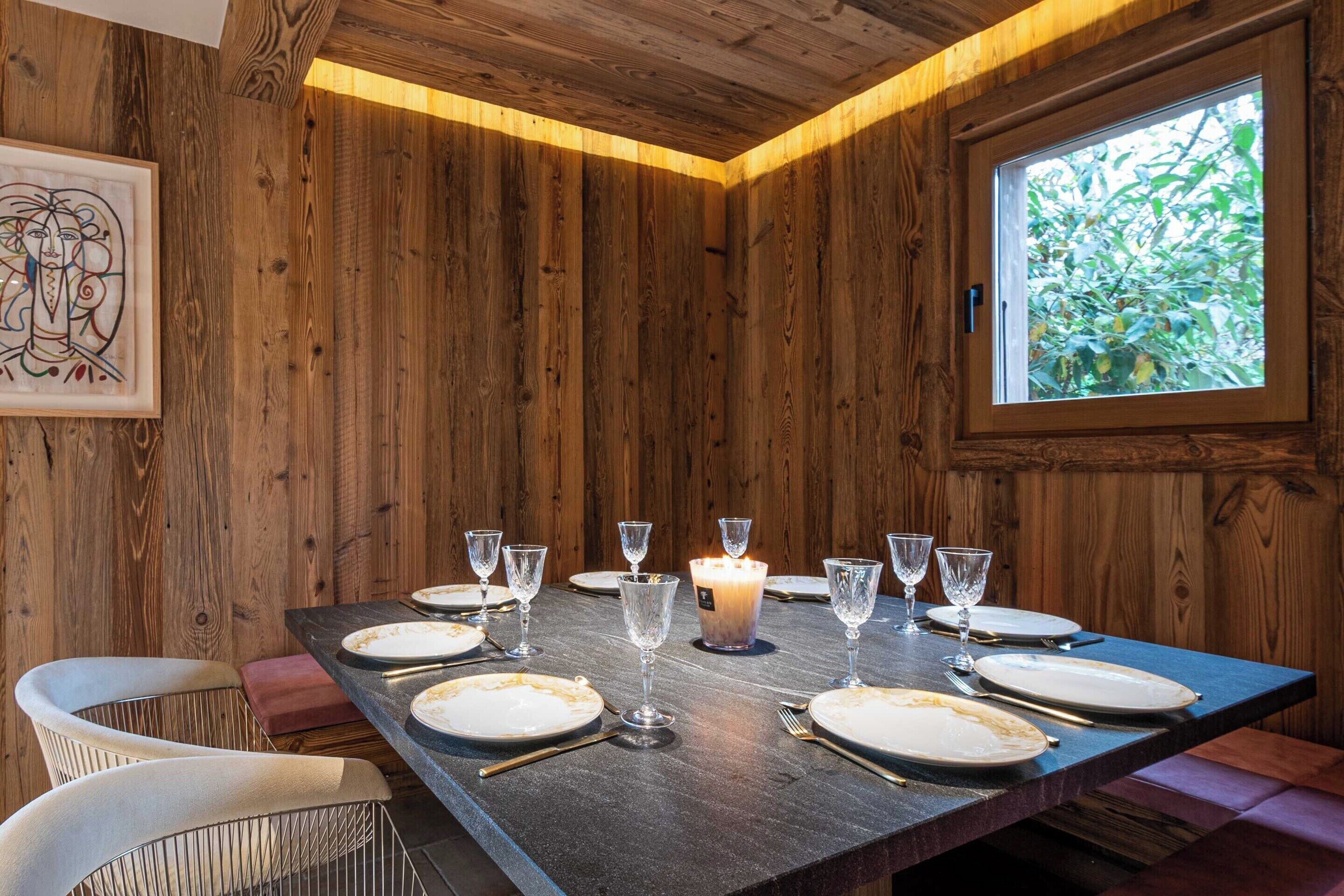 Une pièce aux murs lambrissés, avec une table à manger dressée pour six personnes, une bougie et une fenêtre donnant sur un paysage verdoyant.