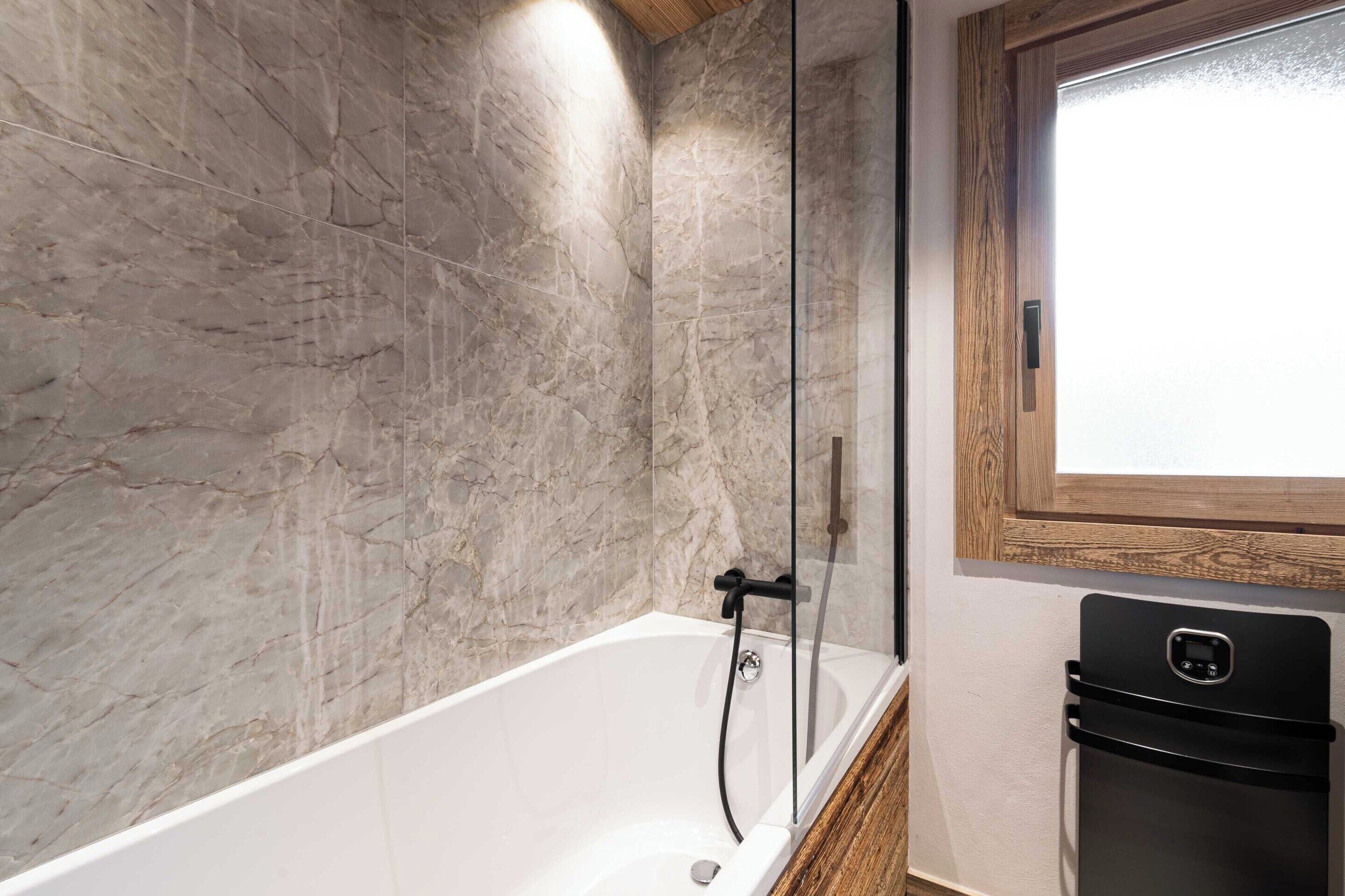A bathroom with a marble wall, a bathtub, and a black refrigerator.