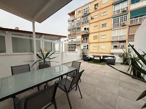 A modern outdoor patio with a glass table and chairs, surrounded by potted plants and a multi-story building with balconies. - Huelin Beach House: Solarium 200 m from the Sea (Malaga)