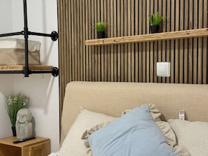 A room with a bed, a wooden shelf with plants, a wall-mounted light switch, and a small wooden table with a vase and a remote control. - Huelin Beach House: Solarium 200 m from the Sea (Malaga)