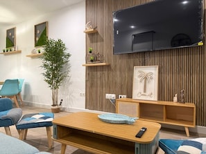 Een moderne woonkamer met een houten tv-meubel, een grote televisie en een gezellige zithoek met blauwe stoelen en een houten tafel.