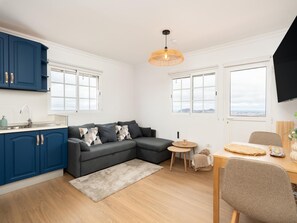 A modern kitchen with blue cabinets, a grey sofa, a small round table, and a TV mounted on the wall. - Vivelorural Casa Marao Great sea Views (Arucas)