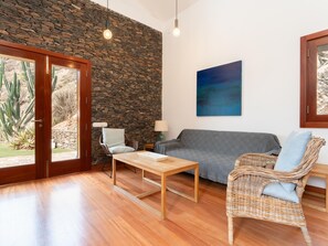 A living room with a stone wall, a grey sofa, a wicker chair, a wooden table, and a painting on the wall. - Vivelorural El Olivo de Ayagaures (San Bartolomé de Tirajana)