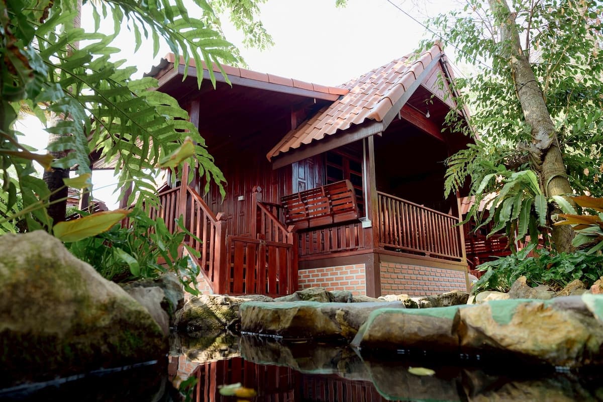 Une cabane en bois dotée d’un balcon, entourée d’une végétation luxuriante et bordée d’un petit étang.
