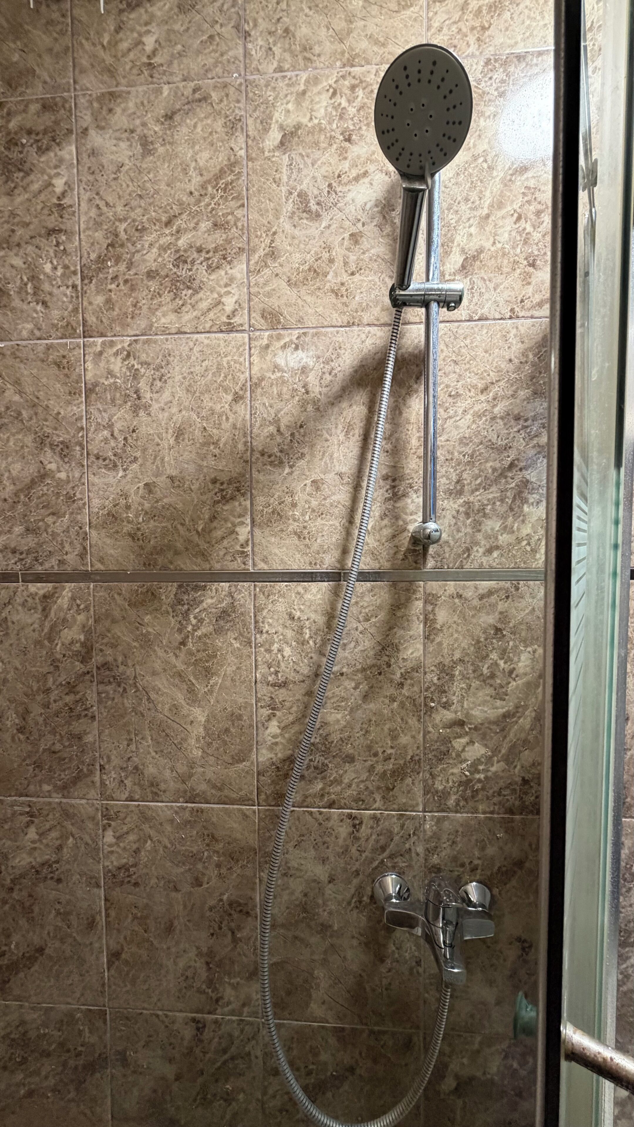 Chuveiro com chuveiro manual e mangueira flexível, fixado numa parede revestida de azulejos.