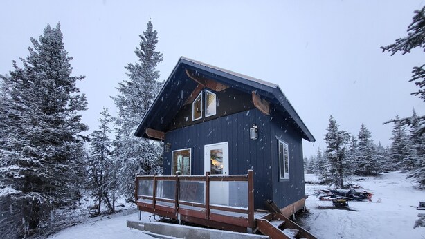 A small cabin with a wooden deck, surrounded by snow-covered trees.