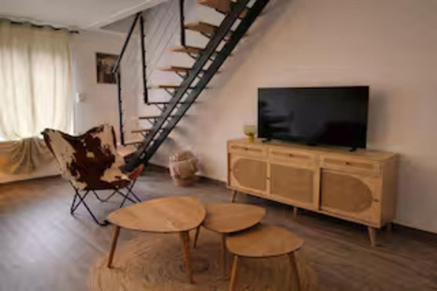 Un salon avec un escalier en bois, un canapé, un téléviseur posé sur un meuble en bois et une petite table ronde.