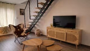 Un salon avec un escalier en bois, un canapé, un téléviseur posé sur un meuble en bois et une petite table ronde.