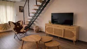Un salon avec un escalier en bois, un canapé, un téléviseur posé sur un meuble en bois et une petite table ronde.
