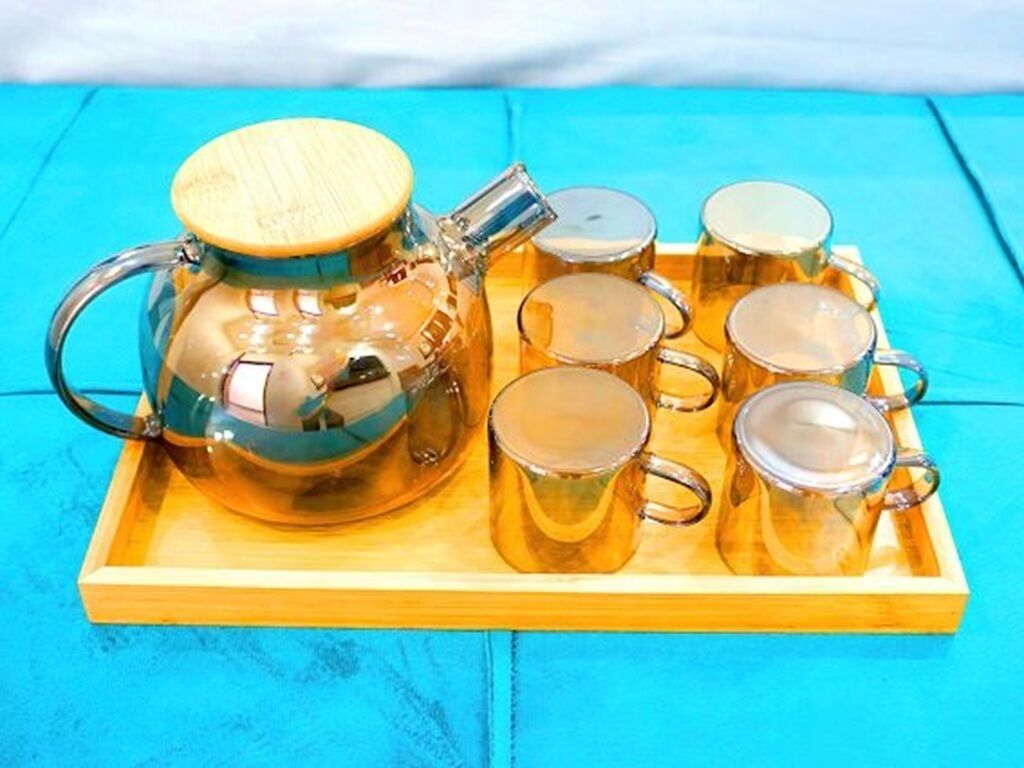 Une théière en verre transparent, dotée d’un couvercle en bois et d’un bec verseur en métal, accompagnée de six tasses en verre assorties, présentées sur un plateau en bois.