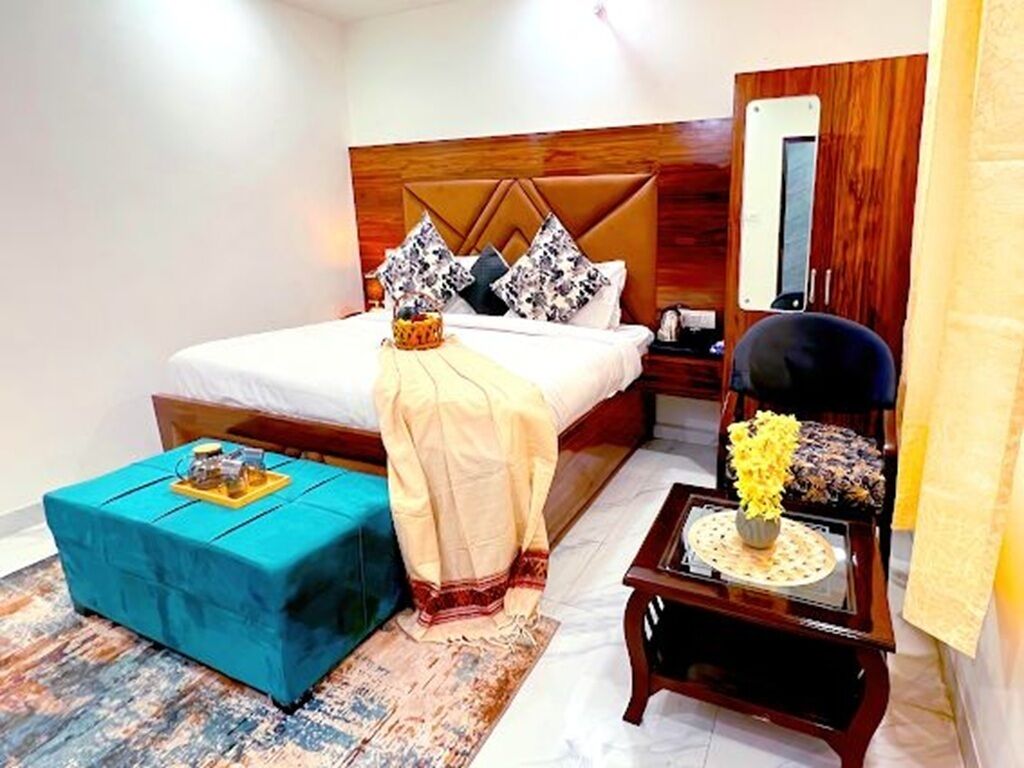 Une chambre à coucher avec un lit, un pouf bleu, une table de chevet en bois, une chaise et une petite table sur laquelle est posé un vase de fleurs.