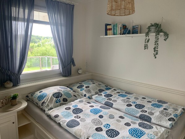 Un dormitorio con dos camas, una ventana con cortinas, una mesita de noche con una planta, una estantería con libros y una planta, y una lámpara colgante.
