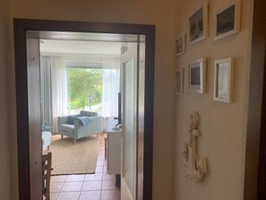 A doorway leading to a living room with a sofa, a television, and a view of greenery outside.