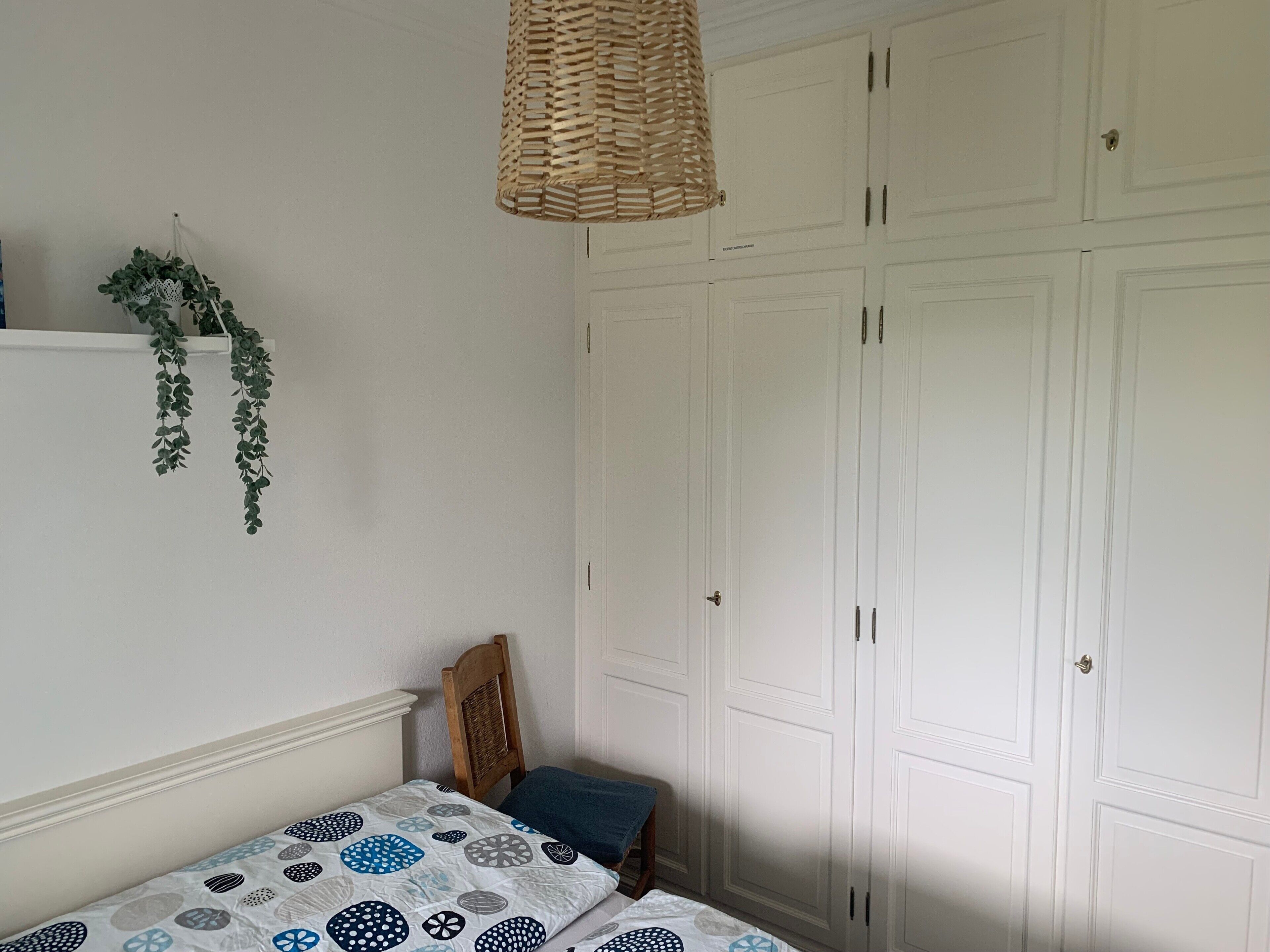 A bedroom with a bed, a wooden chair, a hanging plant, and a wardrobe.