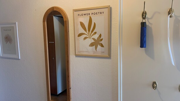 Een kamer met een spiegel met houten lijst, een kunstwerk getiteld 'Flower Poetry' in een lijst, en een deur met een blauwe handdoek aan een haak.
