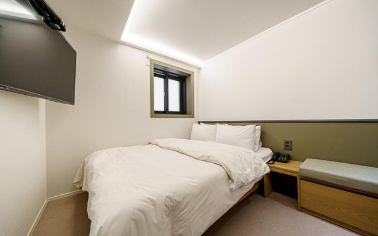 Una cama individual con cabecera, una mesita de noche con lámpara, una ventana pequeña y un televisor fijado a la pared.