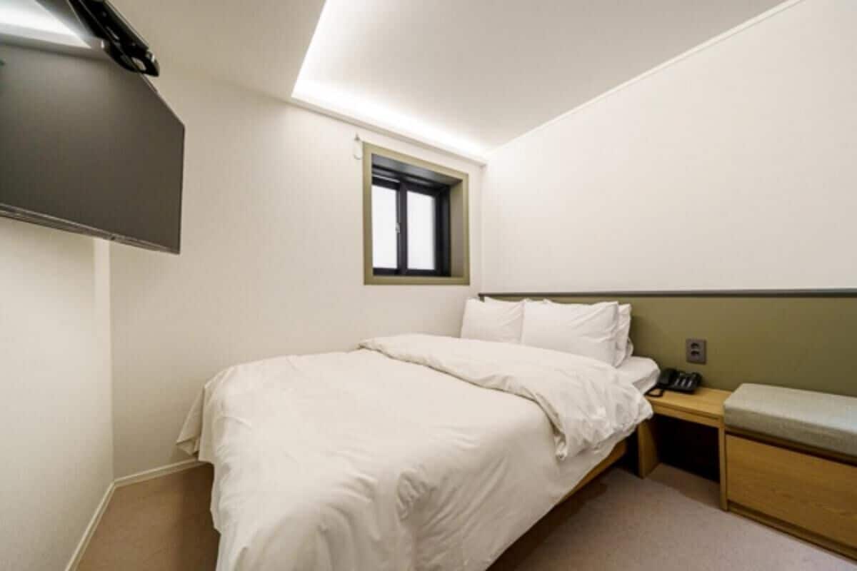 Ein Einzelbett mit Kopfteil, ein Nachttisch mit Lampe, ein kleines Fenster und ein an der Wand befestigtes Fernsehgerät.
