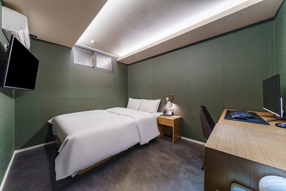 Ein Hotelzimmer mit einem Bett, einem Nachttisch mit Lampe, einem Schreibtisch mit Computer und einem an der Wand befestigten Fernseher.